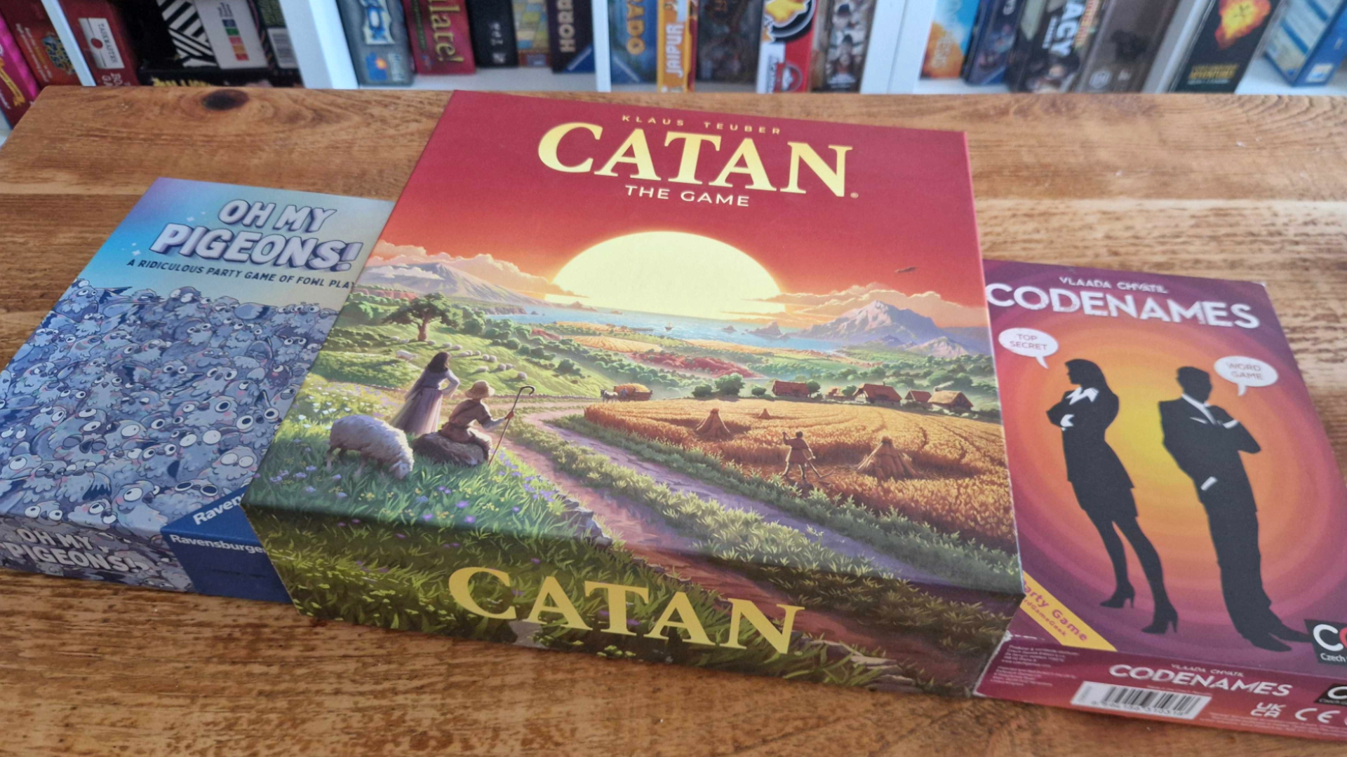 Catan, Oh My Pigeons, and Codenames on a wooden table in front of a board game shelf