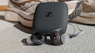 Sennheiser Momentum Sport in-ear headphones in front of charging case, leaning against sports shoe
