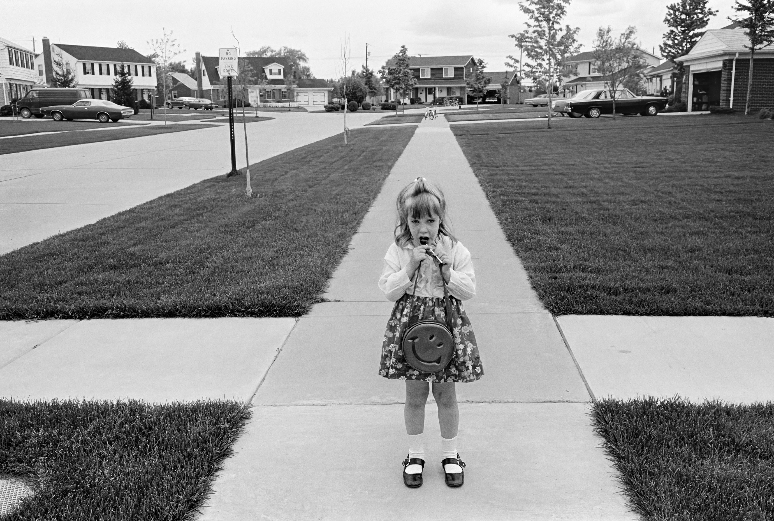 A small girl with a smiley-face purse eats a lollipop as she stands on a suburban pavement lined with neat lawns, young trees and houses.