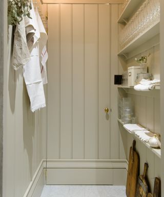 small pantry with shiplap walls painted in stone, shelving with napkins and glassware, chopping boards on the floor, tea towels and linens hanging from hooks on the left