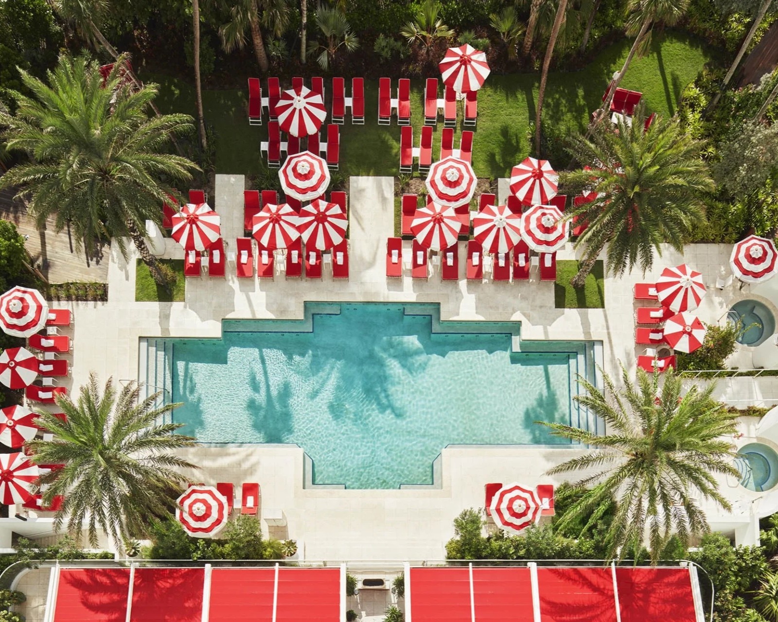A sun-lit pool decorated with stripy red and white parasols and red loungers and palm trees, seen from above.