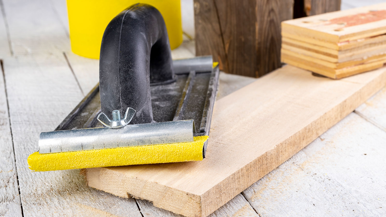 Black manual hand sander with yellow sandpaper lying top of a piece of pine wood on light wooden floor