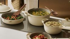 A set of cream colored cookware on a black glass stove top, full of a variety of foods