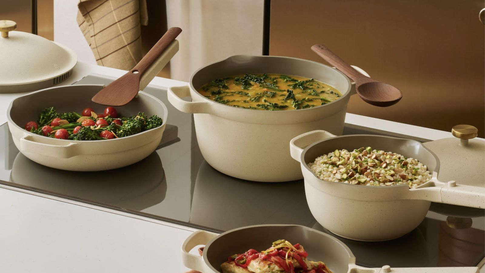 A set of cream colored cookware on a black glass stove top, full of a variety of foods
