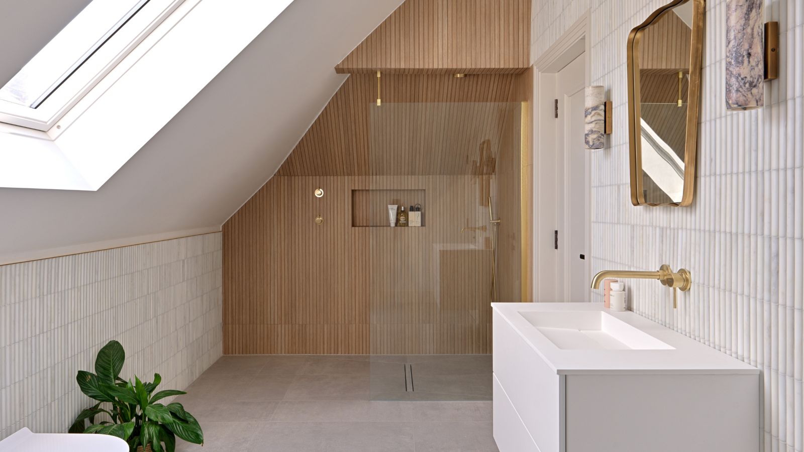 A bright Scandinavian-inspired attic bathroom with a large rooflight and sloped ceiling clad in warm oak slat panelling enclosing a walk-in shower with brass fittings and a recessed niche. Slim white vertical tiles line the lower walls alongside a white floating vanity with a brass wall-mounted tap, gold-framed mirror and a large leafy plant