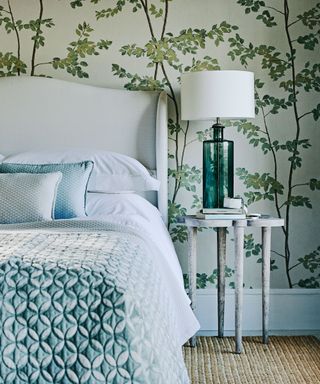 A wall with botanical print wallpaper behind a bed with a white headboard and a light blue throw