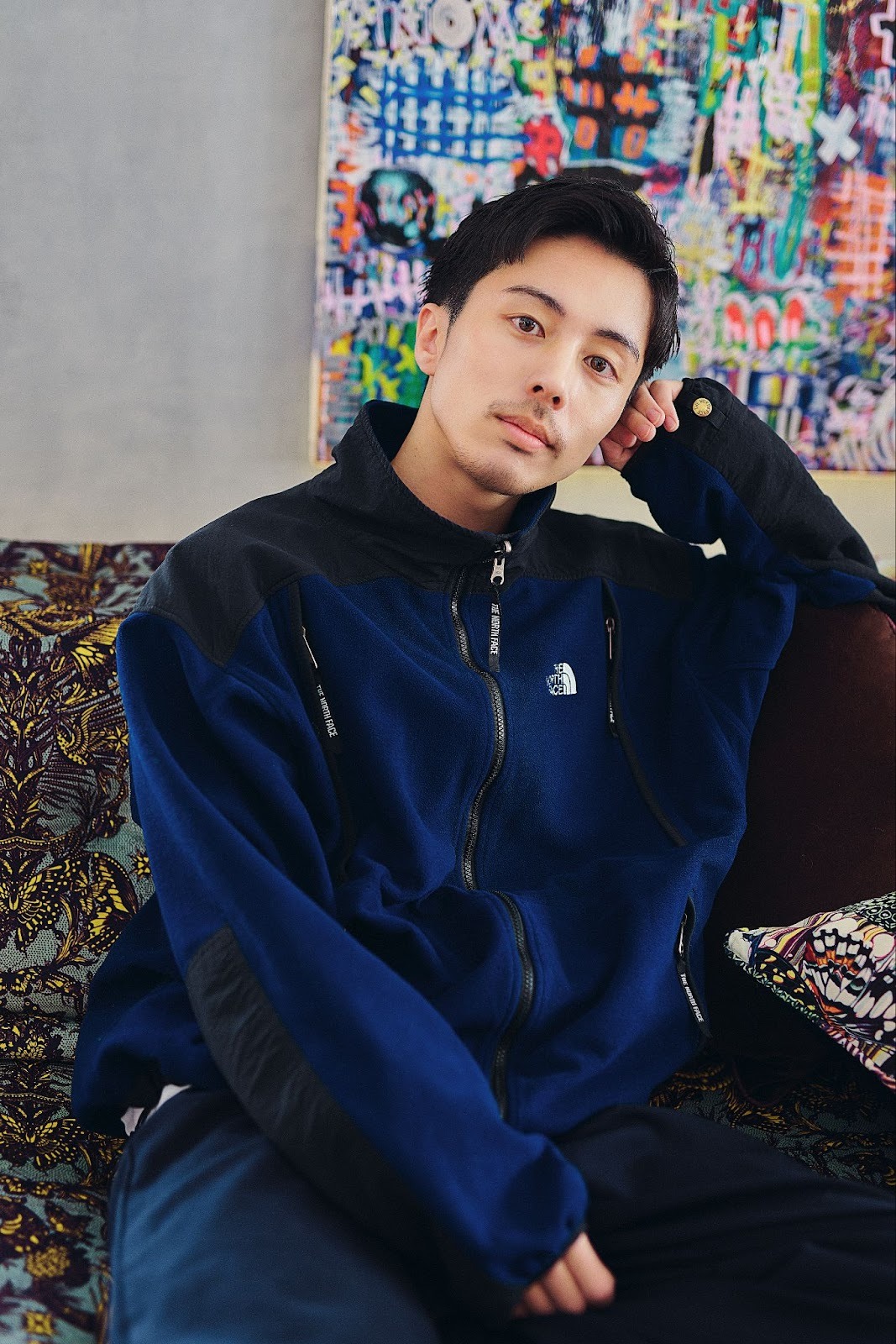 A Japanese man named Hiroya wears a navy winter jacket and navy pants while posing with his head resting on his hand, and sitting on a printed sofa, in a cast portrait for 'The Boyfriend.'