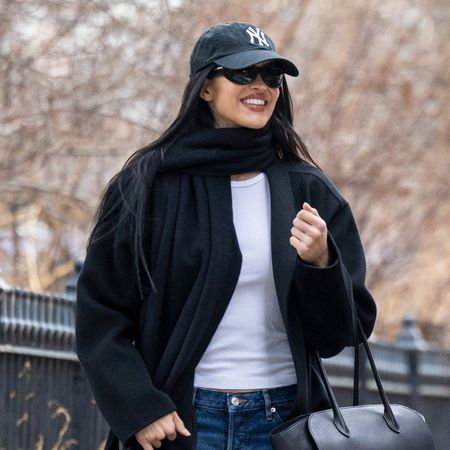 Amelia Gray wearing a Yankees baseball cap, black scarf and coat, white T-shirt, baggy jeans, and black glove flats