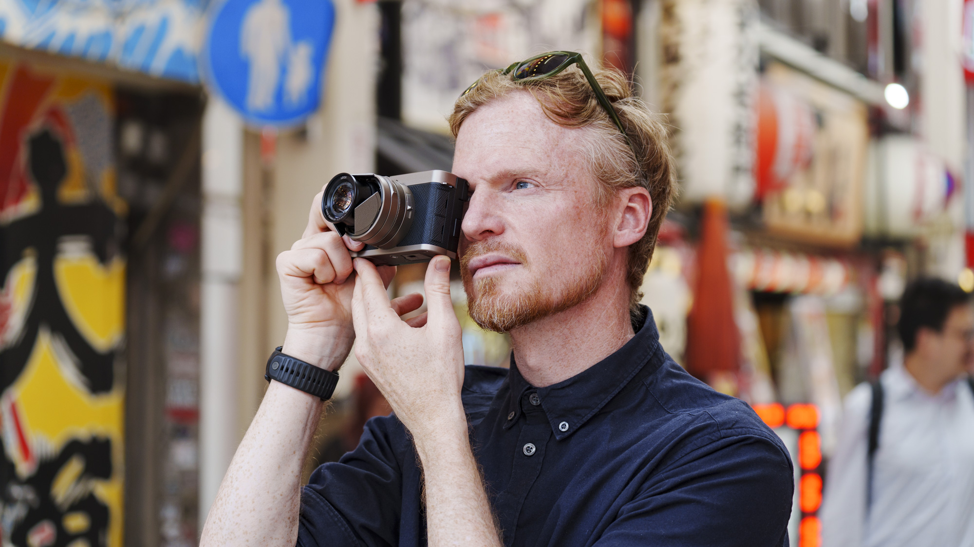 Man holding the Panasonic Lumix L10 compact camera up to his eye, he's looking through the viewfinder, in an urban setting in Japan