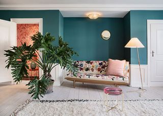 View of a 2013 living area at The Apartment gallery featuring wood flooring, teal walls with white wood panelling at the bottom, a multicoloured floral daybed with a pink cushion, a floor lamp, a round pink and metal side table, a green plant in a pot, a round gold mirror on the wall, a ceiling light, white doors and a white rug. There is a partial view of another space with orange floral walls and a lamp on a side table