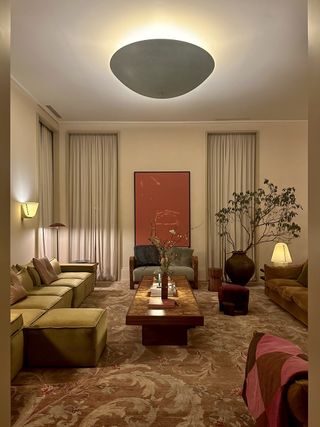 Stylishly furnished concept store, with beige rug, green sofa and a large plant in the corner. On the back wall are two curtained windows with a red painting in the middle