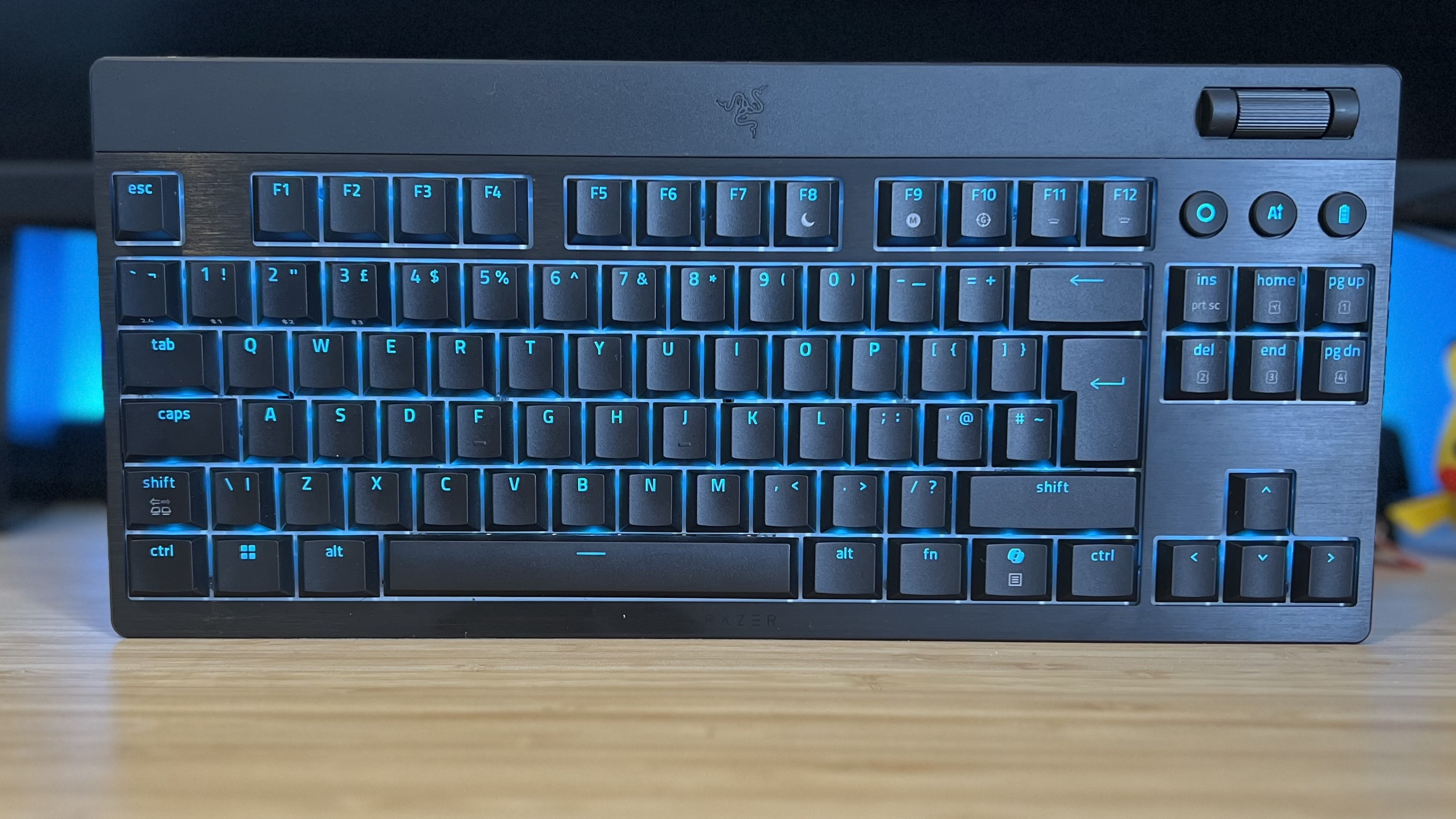 Razer Blackwidow V4 Low Profile TKL gaming keyboard on a wooden desk with blue backlighting