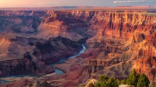 a deep canyon with a river meandering through it. The rock formations are colored a rusty red/orange from the sunset.