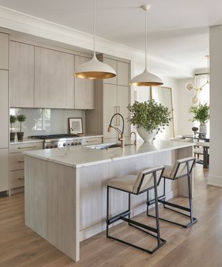 Bright and spacious kitchen with large marble and light stained wood island, light wooden floor, counter chairs with black metal frame and upholstered seat and back cushions, two pendant lights hanging over kitchen island, beige-gray kitchen units, countertops decorated with plants and framed picture, sink on island