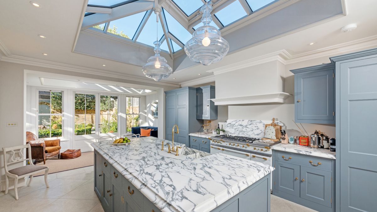 inside of orangery kitchen extension with pale blue kitchen cabinets, marbled worktops and large roof lantern with glass pendant lights hanging above kitchen island