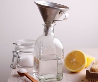 A glass bottle with a stainless steel funnel in the opening with a sliced lemon and teaspoon of baking soda around it