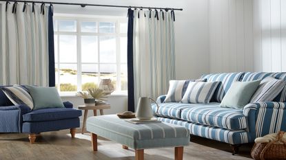 Coastal living room with blue stripe sofa, white linen curtains, striped upholstered coffee table and range of blue pillows and wooden furniture