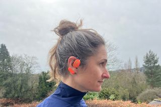 Oladance open ear headphones on a female's ear who is looking to the right side on. There are trees and grey sky in the background
