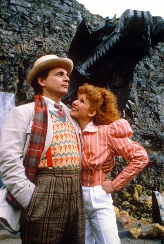 The Seventh Doctor (Sylvester McCoy) stands at the bottom of an imposing cliff. Mel (Bonnie Langford) is leaning against him, smiling and resting her chin on the lapel of his jacket.