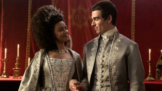 (L to R) India Amarteifio as Young Queen Charlotte, Corey Mylchreest as Young King George holding hands, looking at each other and smiling.