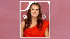 Image of Brooke Shields smiling with curled long brunette hair and wearing a red one-shoulder gown, on a plum background