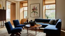 A modern living room with a dark blue corner sofa and two matching armchairs with a round wooden coffee table in front of two inset wood-clad window frames that match the internal doorway
