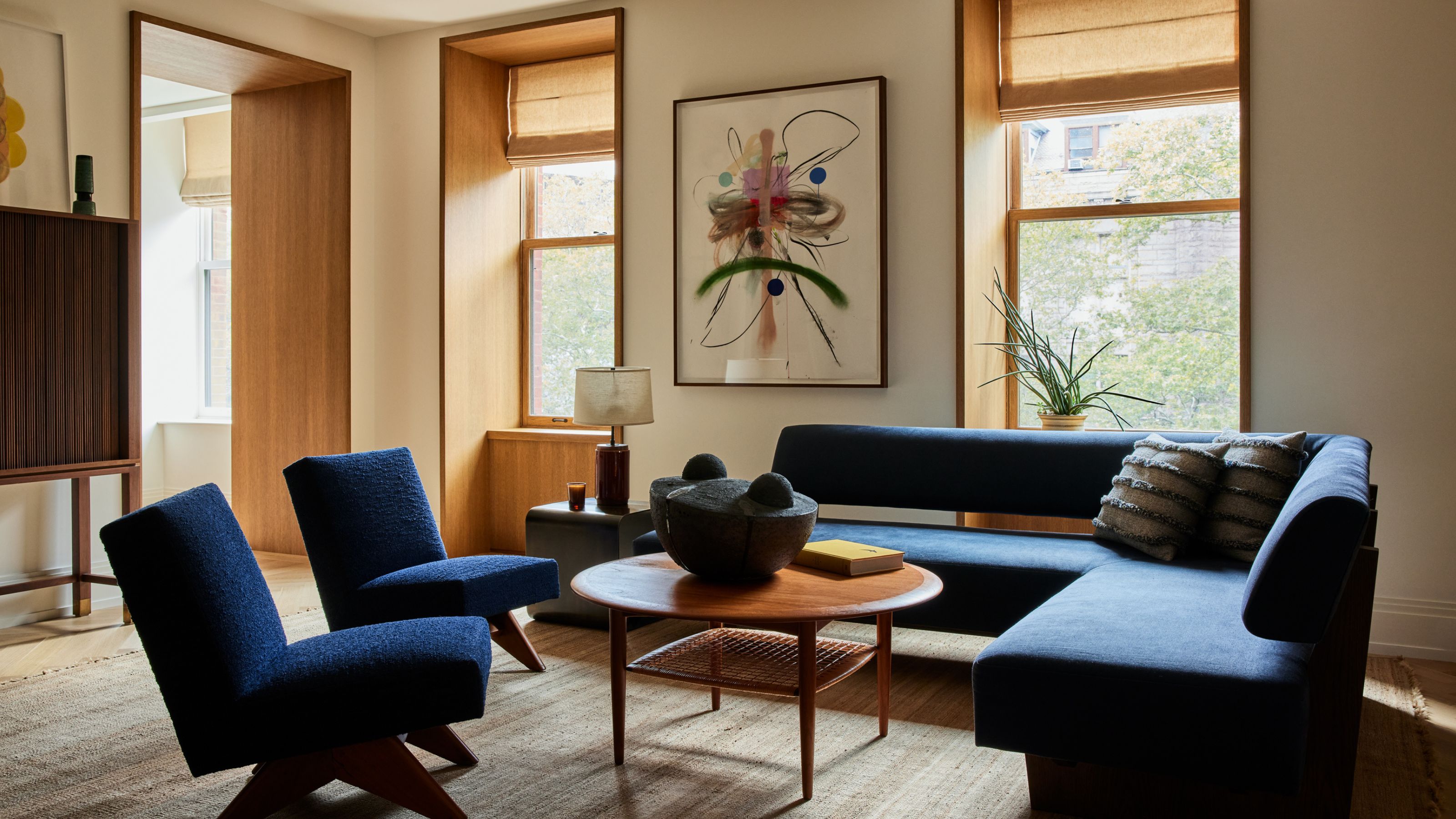 A modern living room with a dark blue corner sofa and two matching armchairs with a round wooden coffee table in front of two inset wood-clad window frames that match the internal doorway