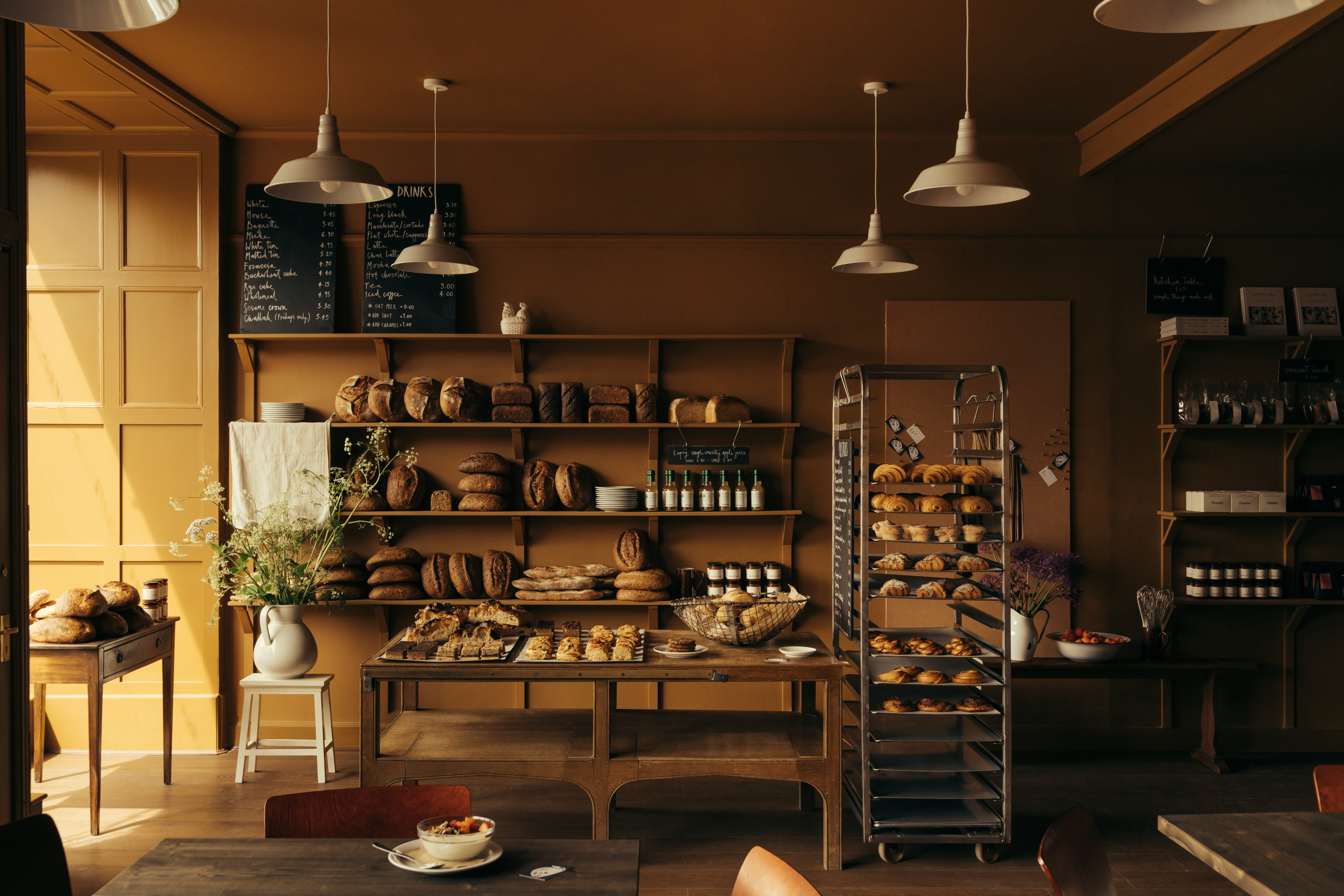 An ochre-painted cafe and bakery filled with wooden furnishings and industrial pendant lighting, along with lots of freshly baked goods, beautifully plated food, and coffee cups.