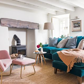 Living room with white walls and an exposed beam above a fireplace, with a large brown rug underneath a green sofa and pink armchair with matching footstool