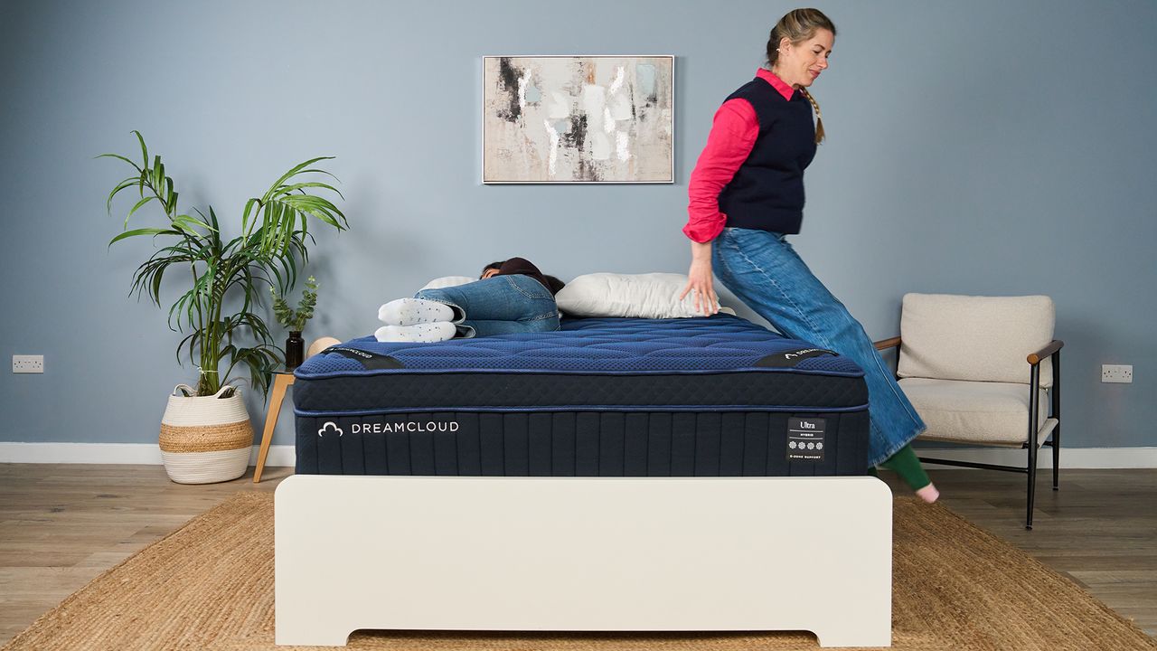 The image shows a mattress tester lowering herself onto a navy blue mattress while another tester lies on it on her side