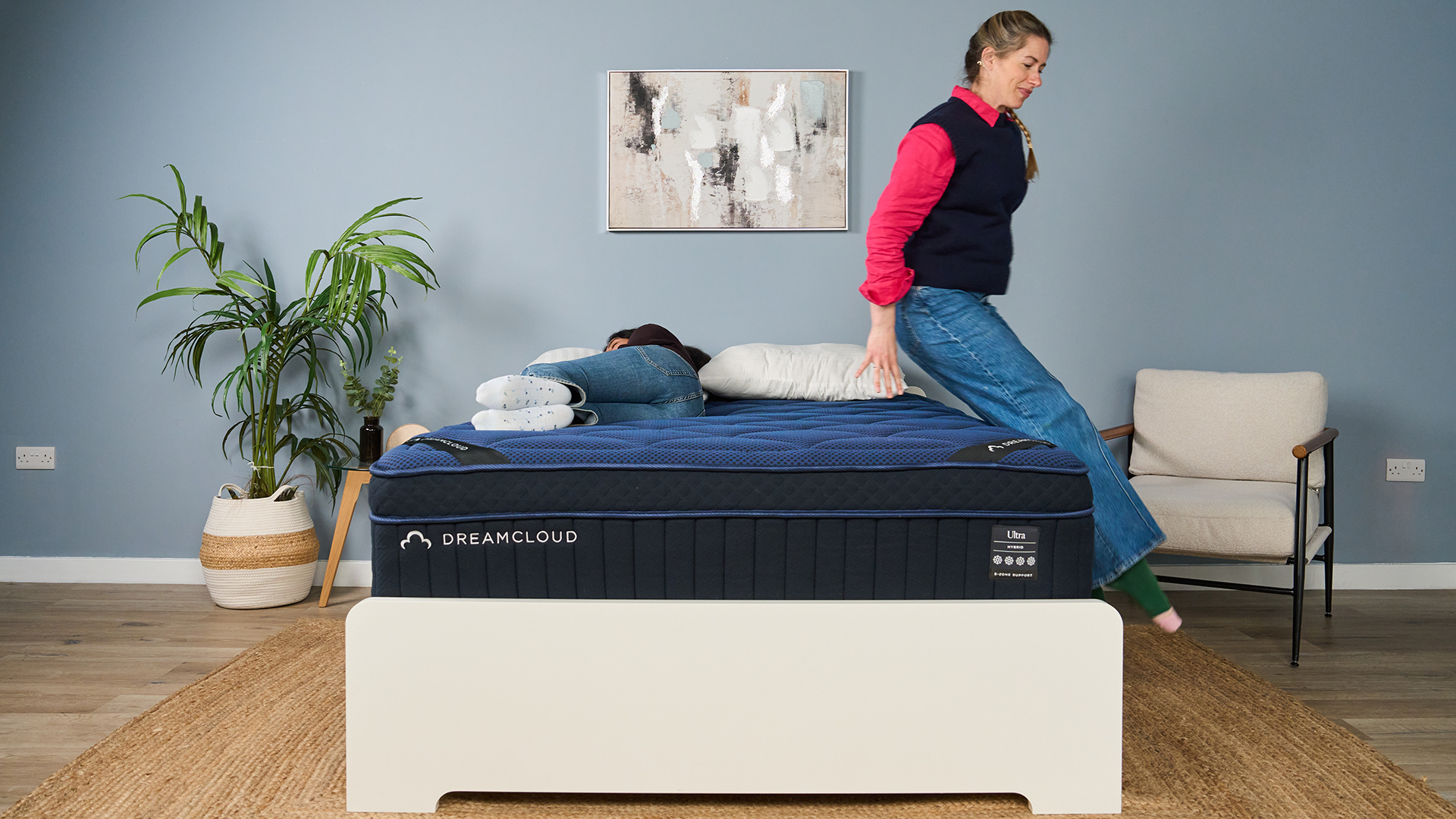 The image shows a mattress tester lowering herself onto a navy blue mattress while another tester lies on it on her side