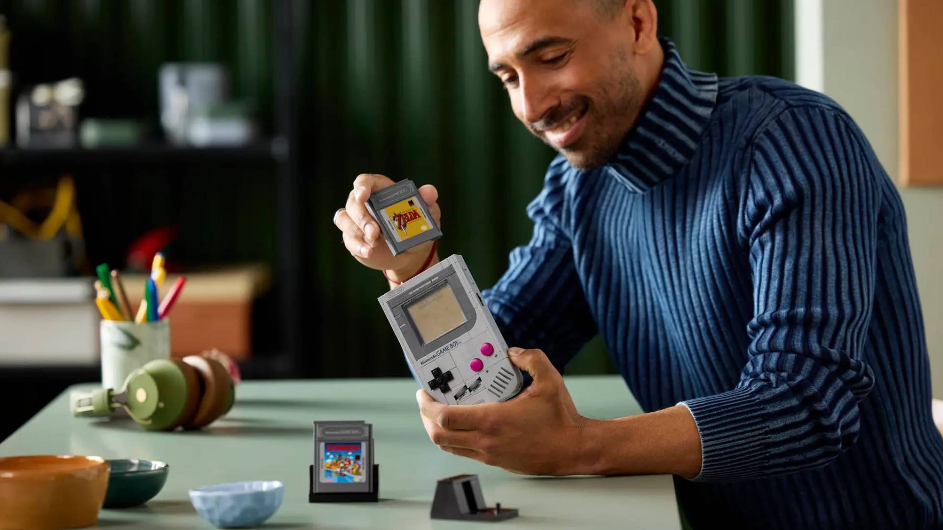 Masculine presenting person with blue shirt holding Lego Game Boy in left hand and Zelda cart in right at desk.