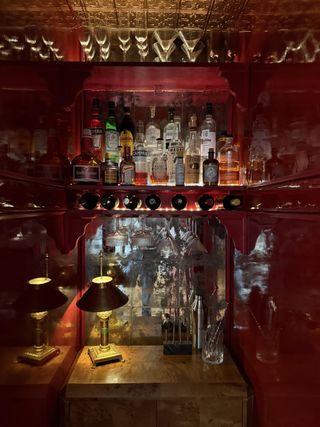 Close-up image of a home bar that is red on the inside. There is a wooden cabinet and a mirrored back wall. On top of the cabinet is a small gold lamp with a black shade and there are bottles and glasses decorating the space