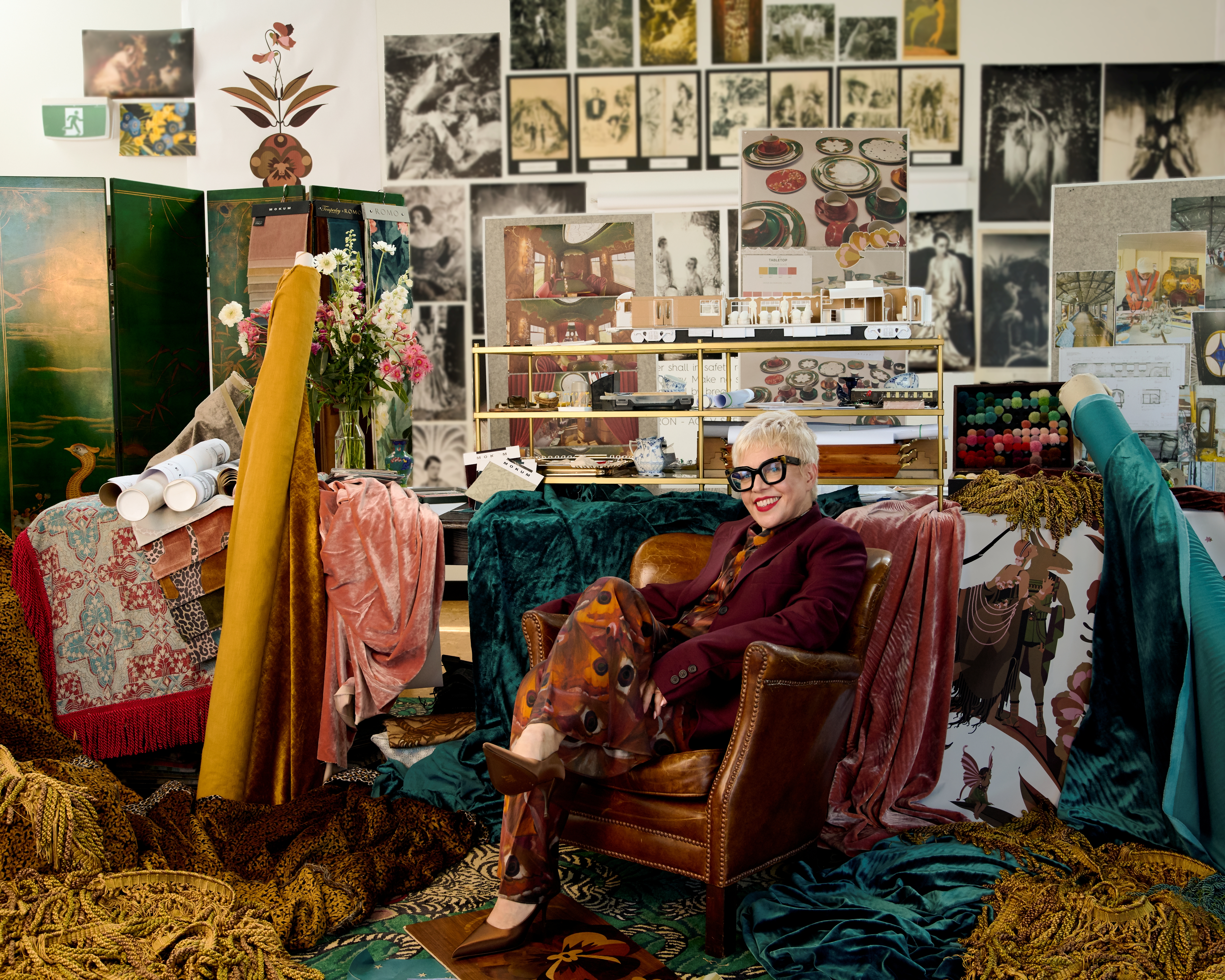 A middle-aged woman dressed in a bordeaux tailored set with brown heels sits while wearing black square glasses on a leathery armchair in a busy, sketches and textiles-filled studio.