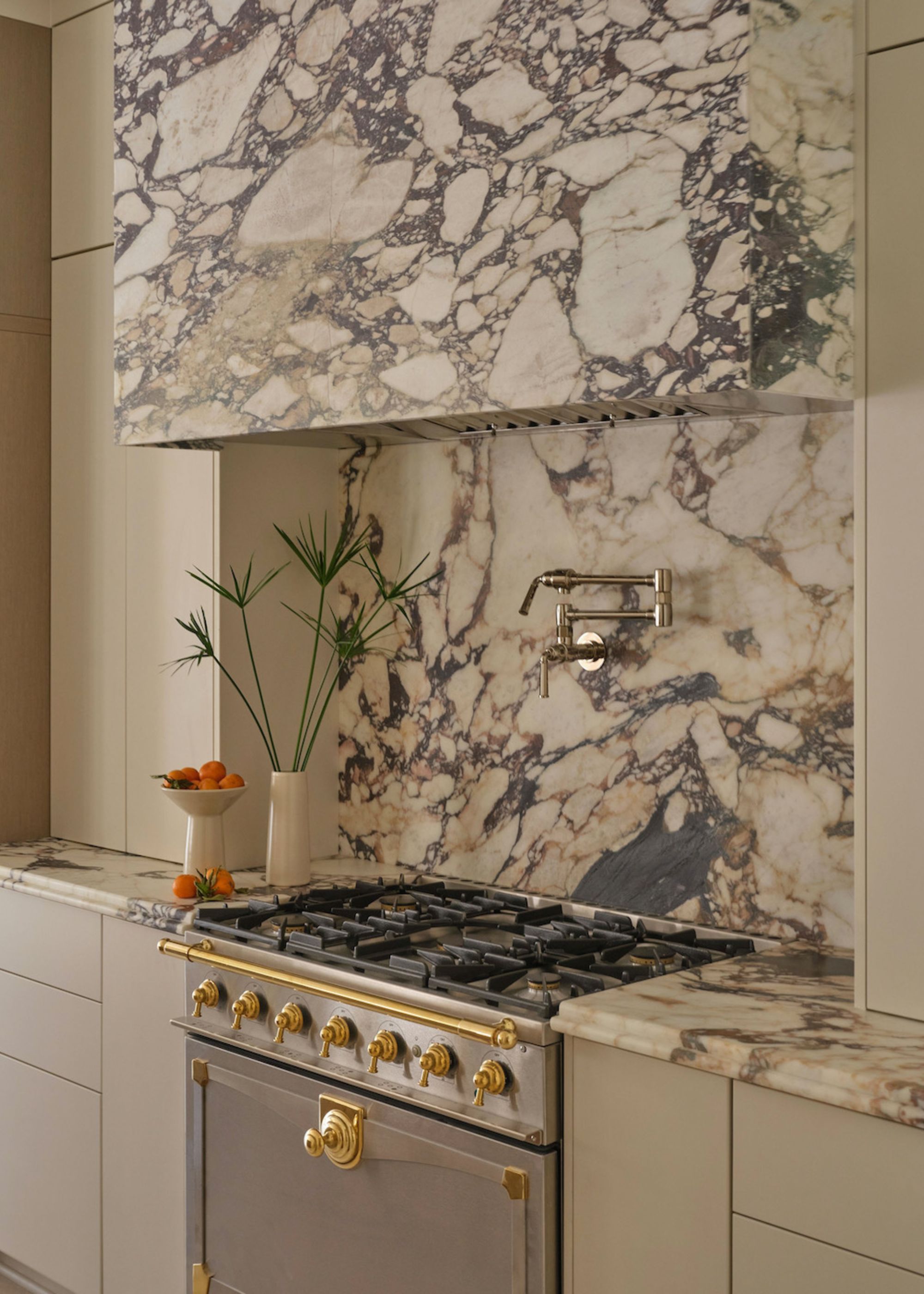 A beautiful richly veined calacatta viola marble extractor hood and backsplash with gold handle and knobs on the oven and a vase of foliage and a bowl of fruit to the left of the oven