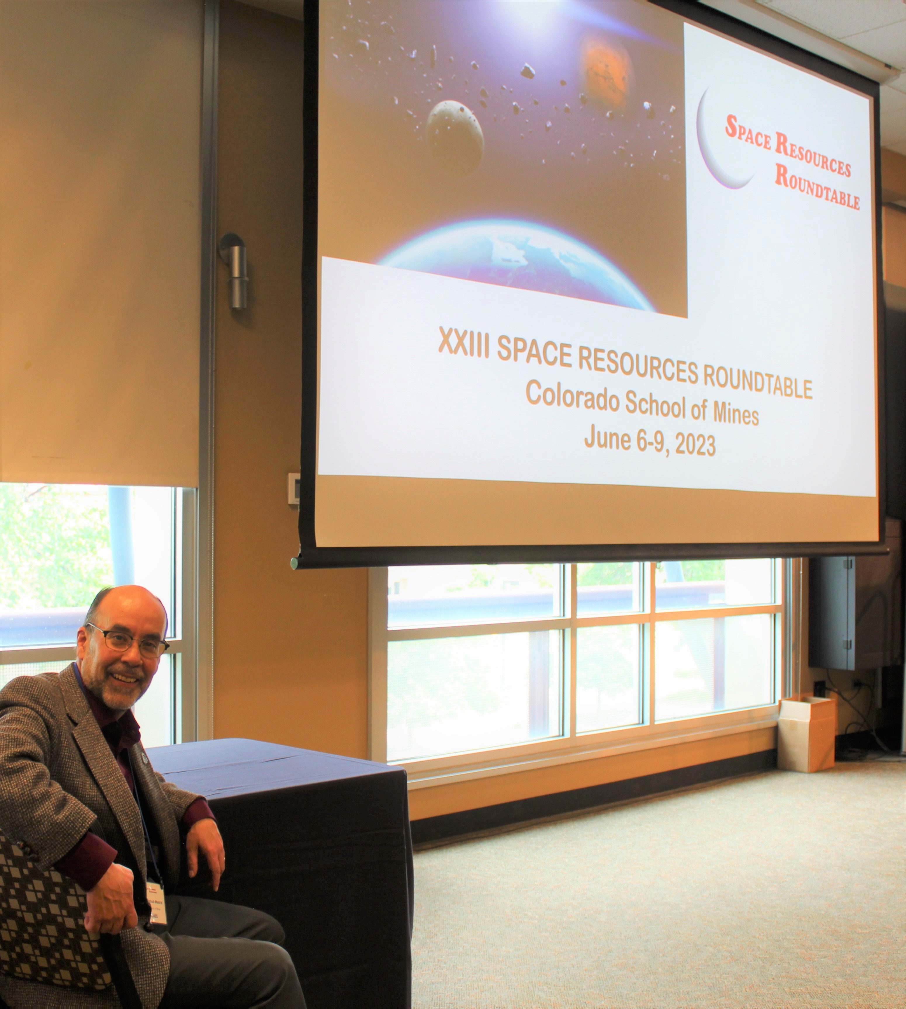 A man sits to the side of a projection screen featuring a space resources slide.