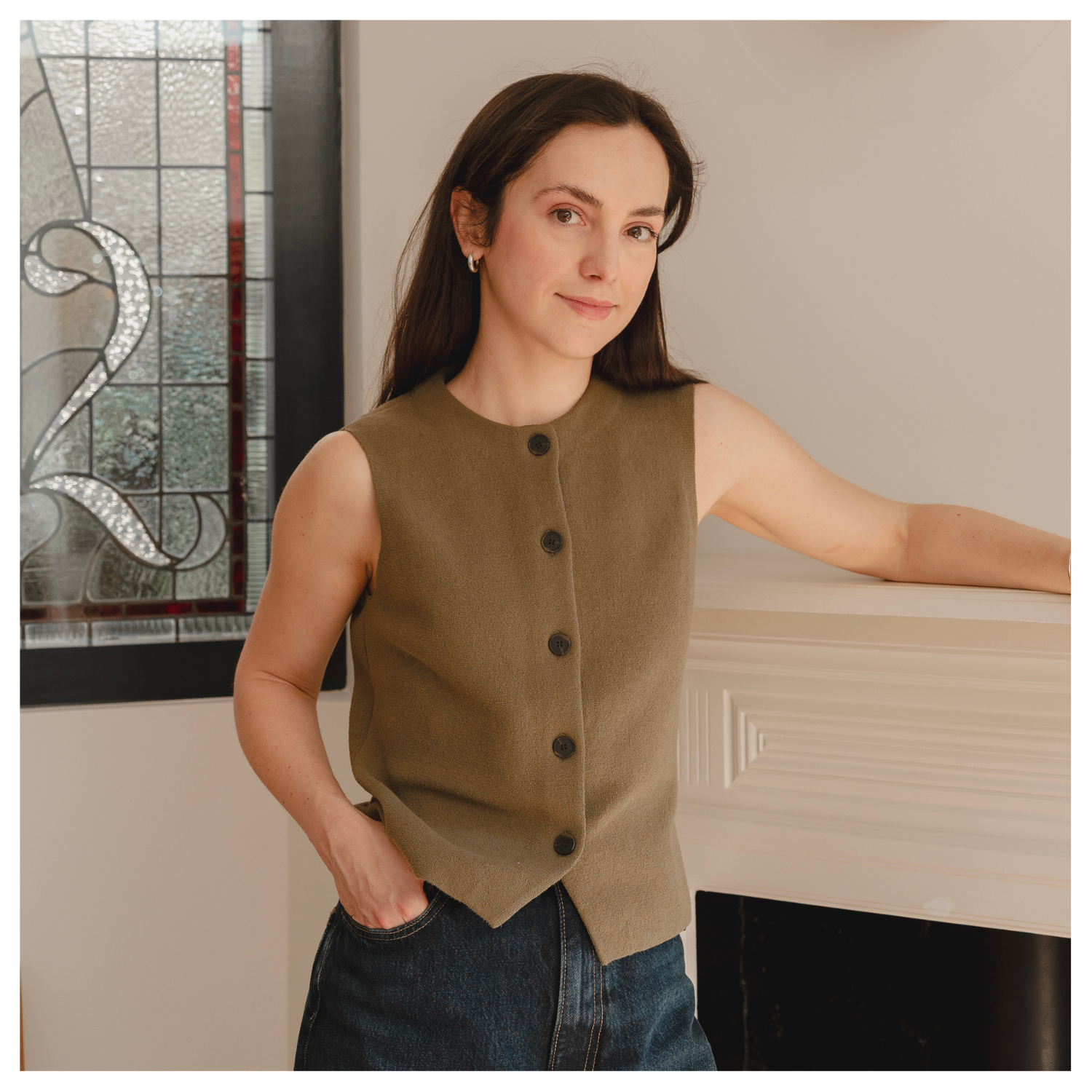 A headshot of Aleksandra Cannock in a buttoned vest and jeans, leaning against a mantel