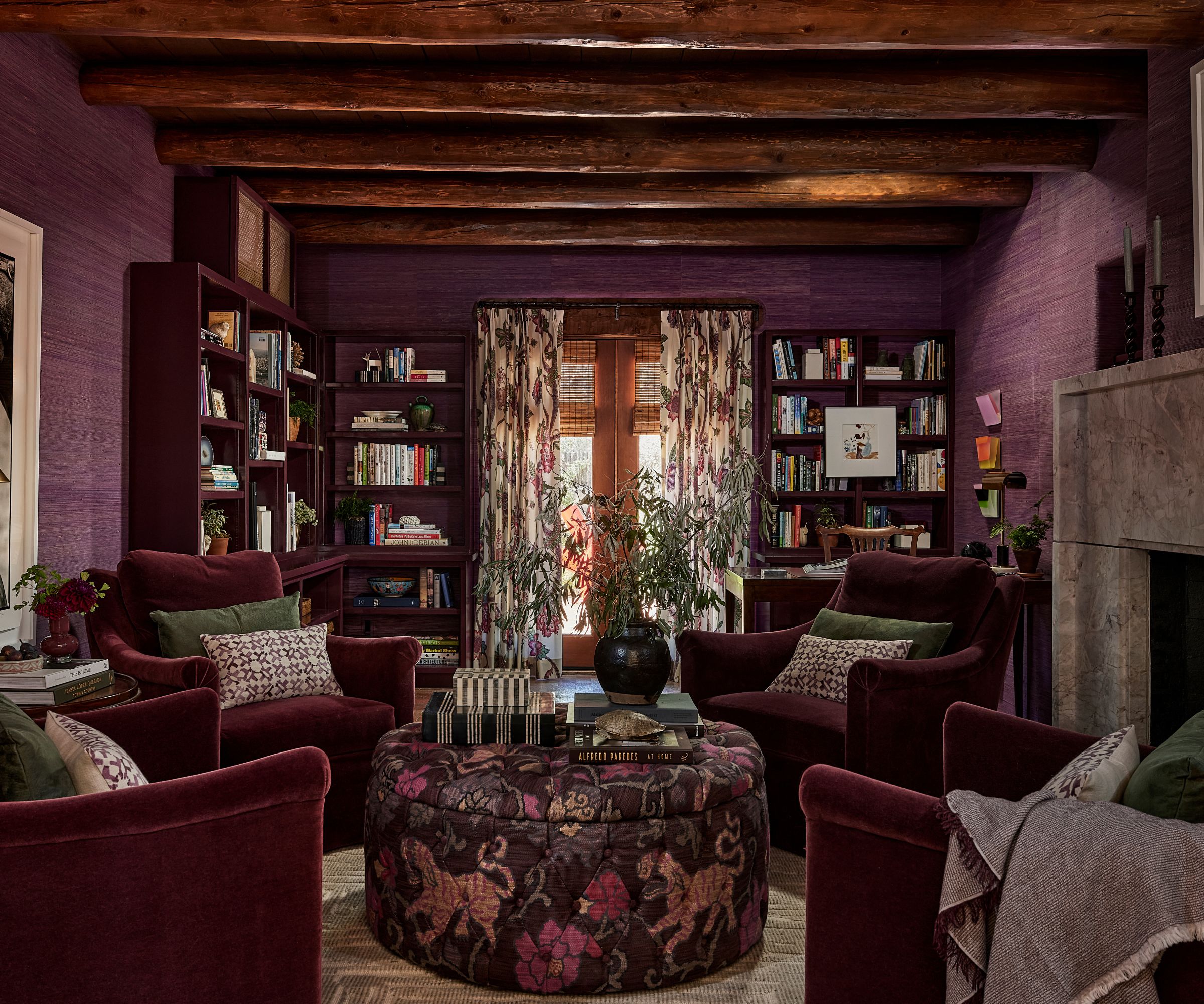 Moody living room with purple grasscloth walls, beamed ceiling, floral drapes, dark red bookshelves, and four dark red armchairs around a floral ottoman with decor on top