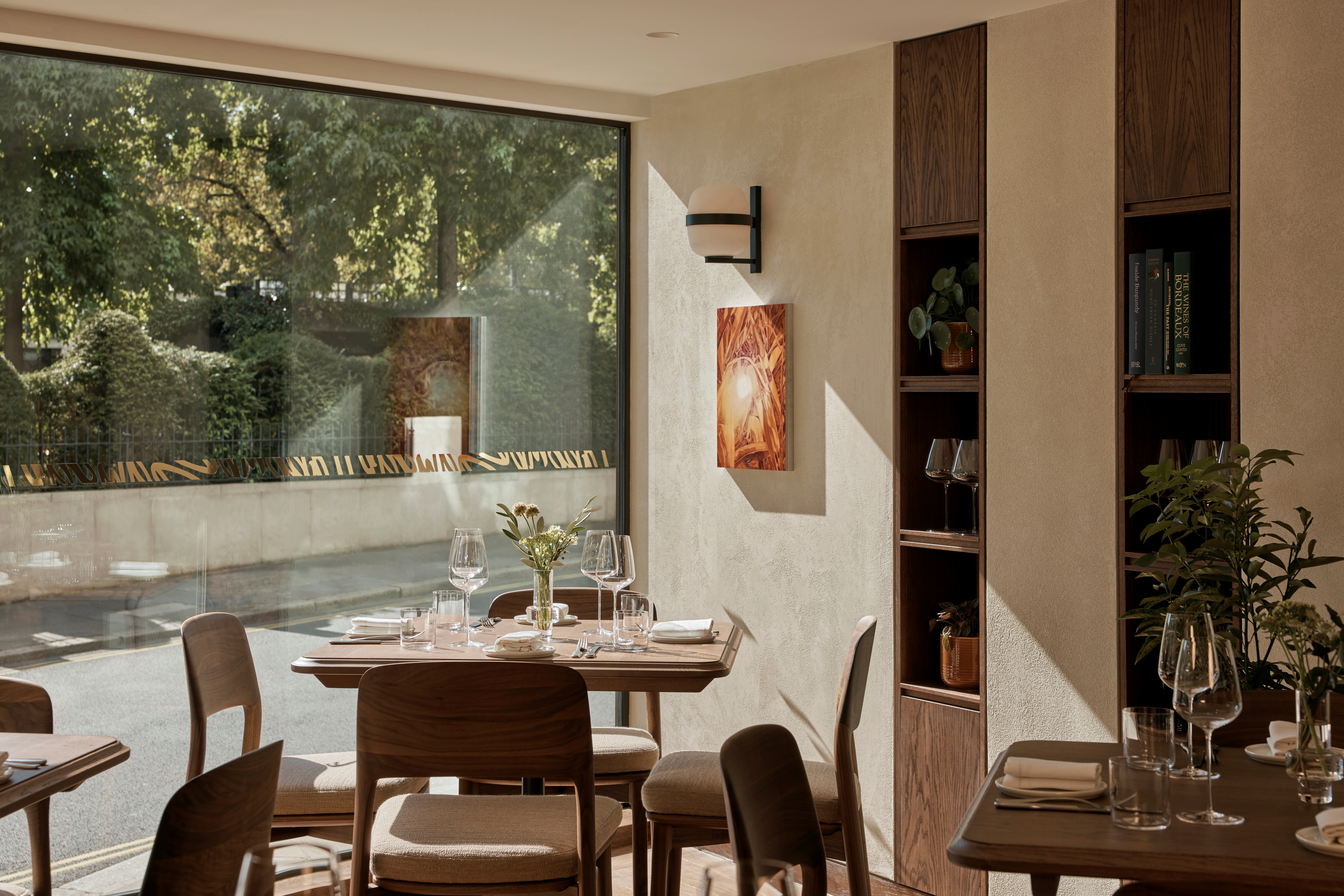 A naturally lit, sunny restaurant with wooden, Scandinavian-style furnishings, shelving filled with glasses, pots, and books, thriving plants, and hung artwork.