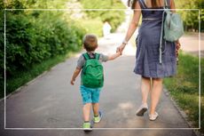 Mother holding hands with child as they walk down a path on a summer's day with backpacks on their backs