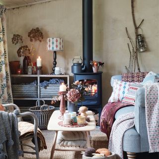 A cosy living room, a blue sofa and chair, basket and quilts and throws. A lit woodburner and kettle, shelves and carpet
