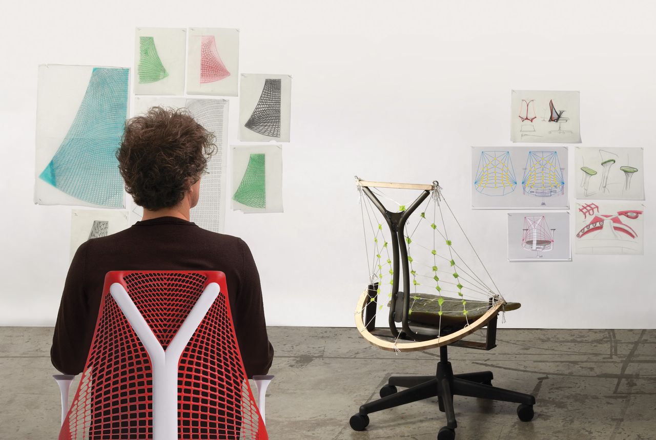 A man sitting on a chair backing the camera and facing a white wall with sketches pinned on the wall