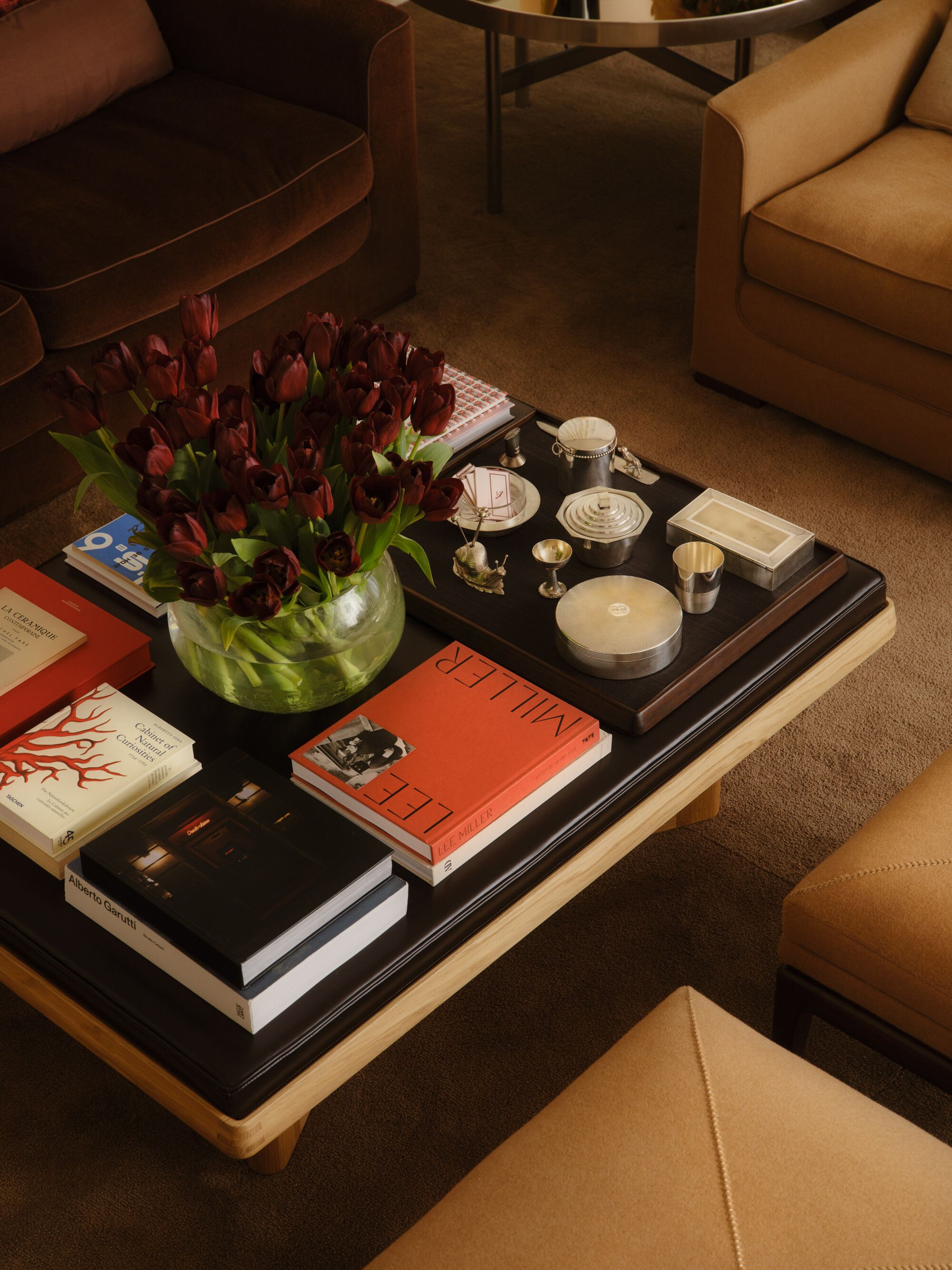 Close up of a black coffee table with lots of coffee table books styled on it, a large vase of red flowers, and some decorative objects. The carpet is tan.