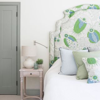 an upholstered headboard with a cream, blue and green flower pattern with matching cushions on the bed, next to a small pink nightstand, next to a grey door