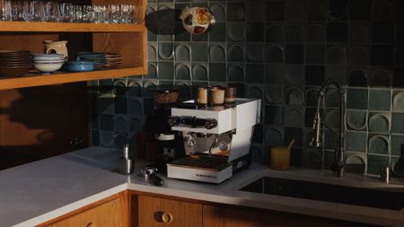 Image of a white La Marzocco home appliance on a white countertop in a kitchen that has wooden cabinetry and sage green backsplash tiles.