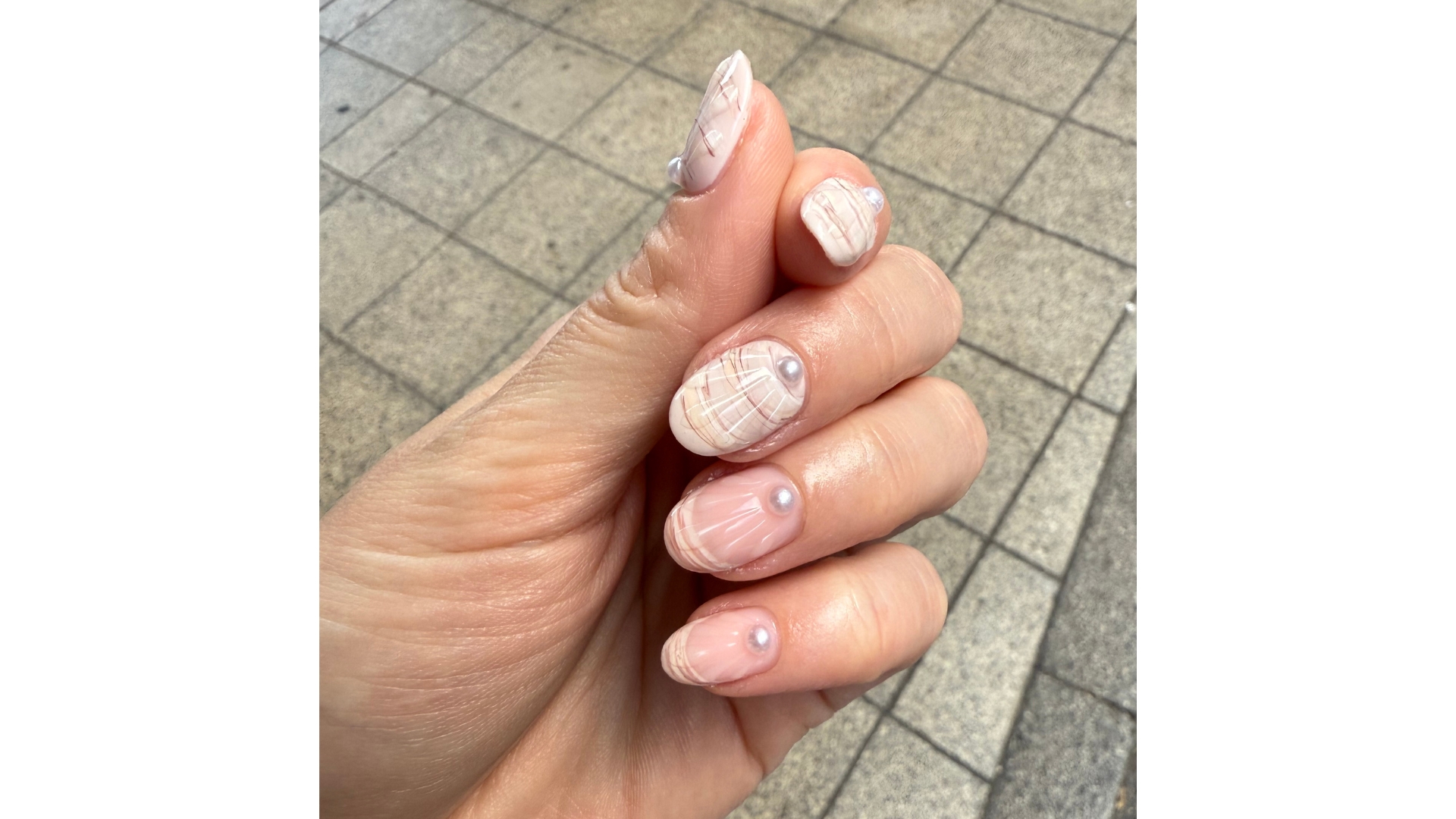 A close-up picture of Contributing Beauty Editor Lucy Abbersteen's hand, which features an almond-shaped manicure pink and white 3D shell nail art, with pearl decals/ taken in front of a pavement background