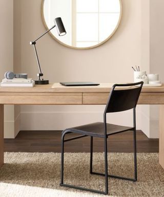 A home office space with a beige wall with a circular mirror, a wooden desk with a laptop and black desk lamp, a black metal chair, and a light gray rug underneath it on dark wooden flooring