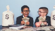 two kids dressed as businessmen holding cash and bookended by white bags with a dollar sign on them