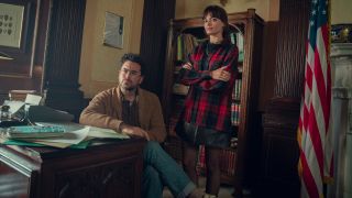 A press image of Emma Mackey standing and Dan Levy sitting behind a desk in Sex Education.