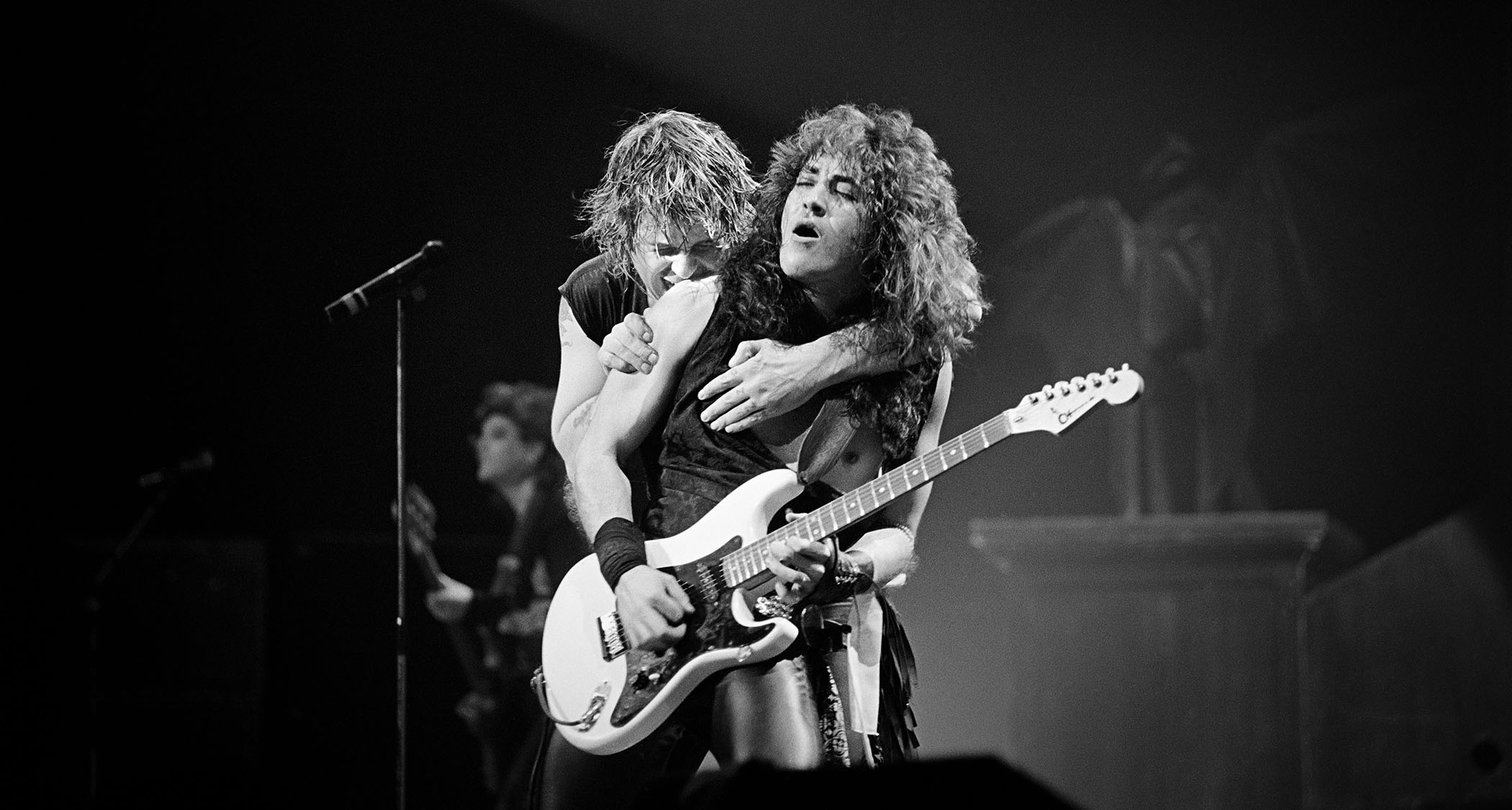 An iconic B/W shot of Ozzy Osbourne embracing Jake E. Lee as he takes a solo during a show.